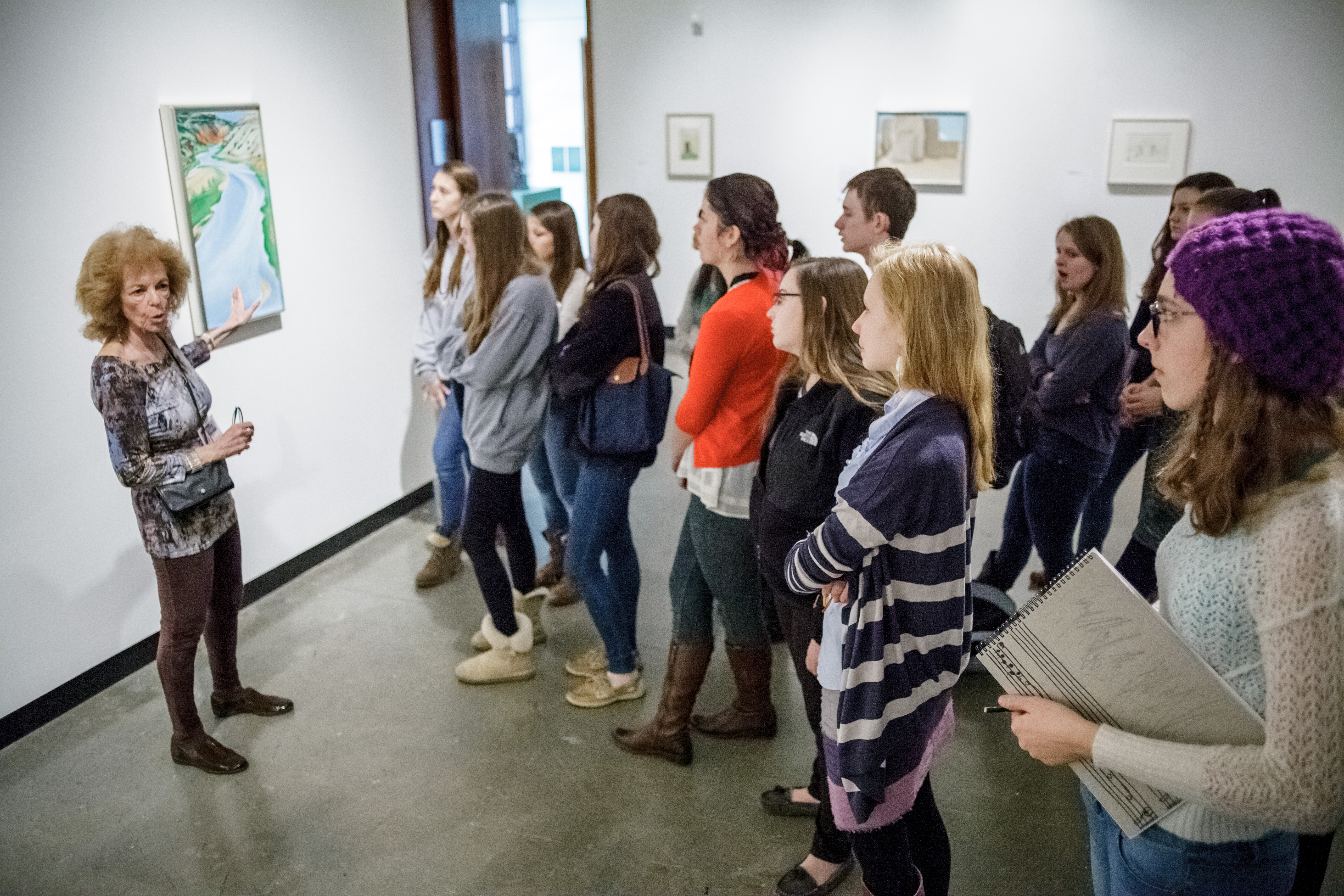 A mam docent is giving a group of high school students a tour in the MAM galleries. The docent is pointing at a painting of a winding road. The students are all very focused on the painting.
