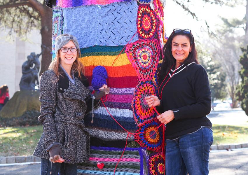 Two volunteers posing next to a large, wide tree trunk that they just decorated with knitted pieces during the MAM Yarn Bomb.