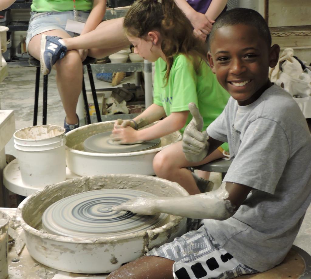 A young student is smiling at the camera and sitting at a pottery wheel. He is covered in clay up to his elbows and looks pretty happy about it. 
