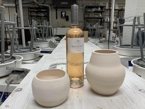 a wine bottle and two handmade pieces of pottery sit in the center table between the rows of pottery wheels in the ceramics studio at MAM.