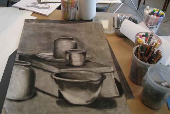 A student's workspace on a table in the studio from the view of the student. A Large sketchbook is in front of us, with a charcoal drawing of some bowls and bottles. To the right is a couple of cups full of pencils and other drawing utensils.