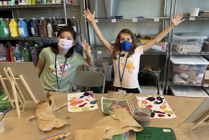 Two young students posing for the camera during painting camp with materials strewn in front of them.