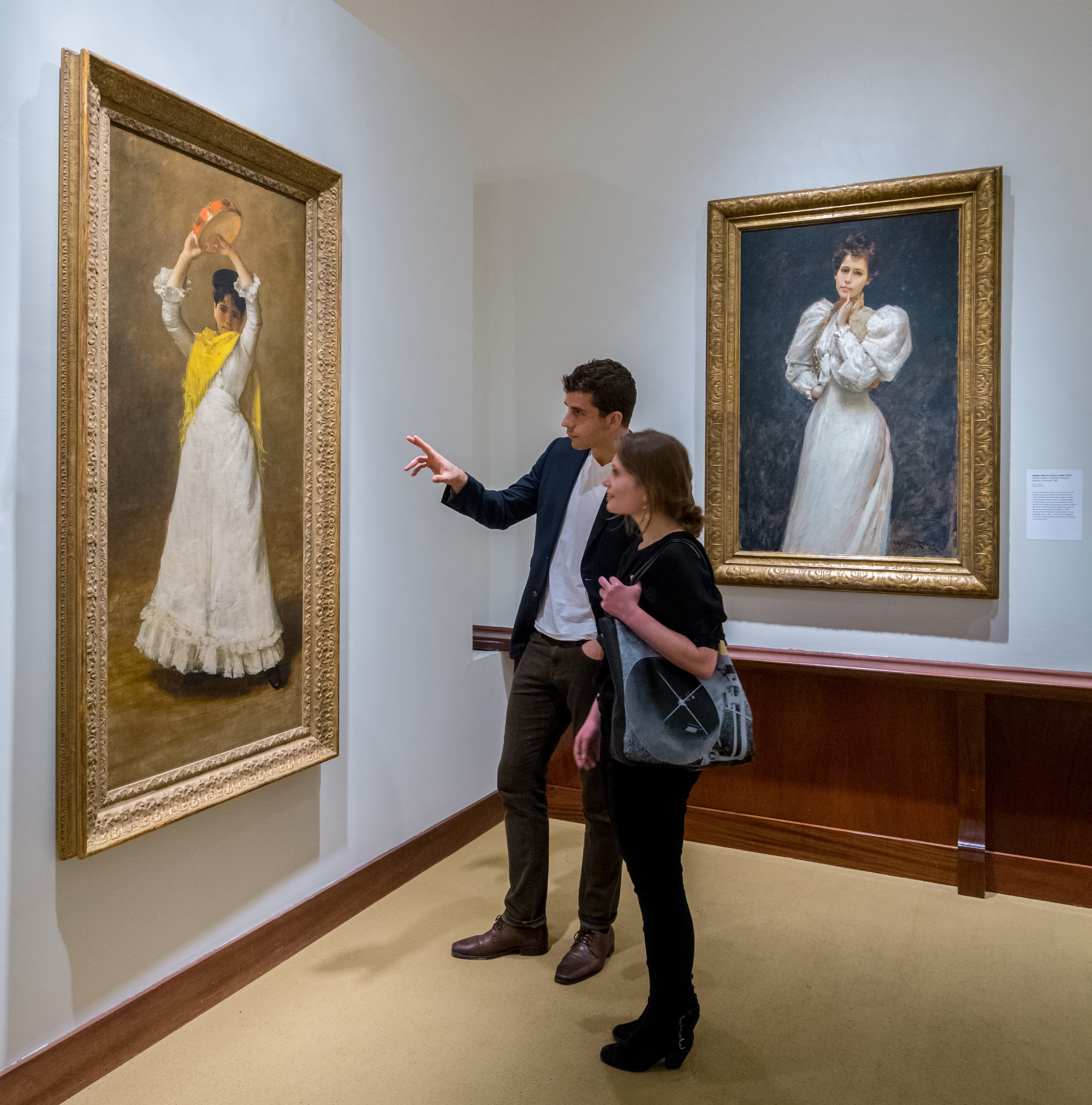 2 visitors in the Marion Mann Roberts Gallery. One man and one women viewed from the side. The man is a head taller than the women and he is pointing to a painting on the wall