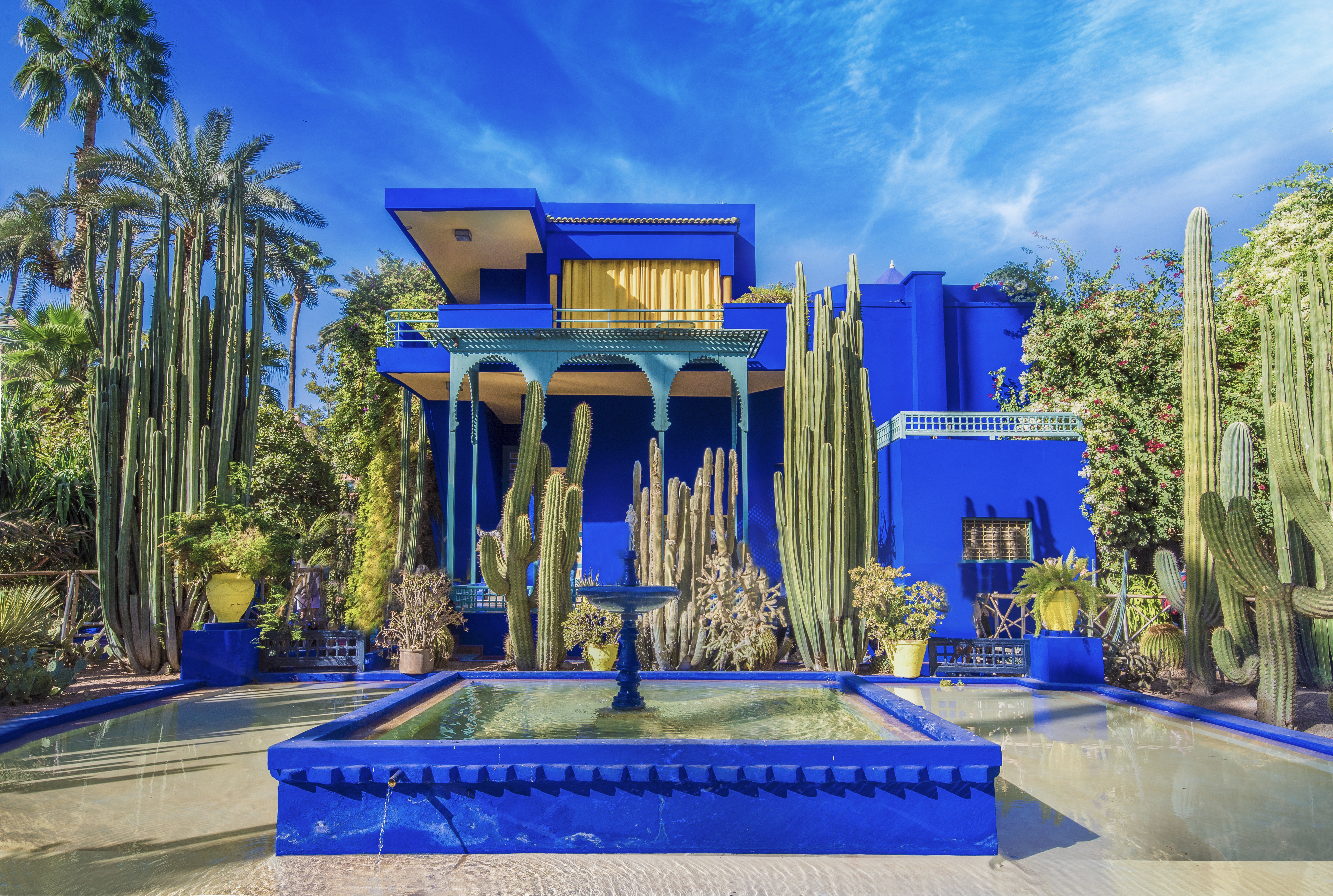 This image is of the Majorelle Garden and Yves Saint Laurant home in Marrakech, Morocco.  The home is a vibrant blue and features a large, square fountain in front of it. 