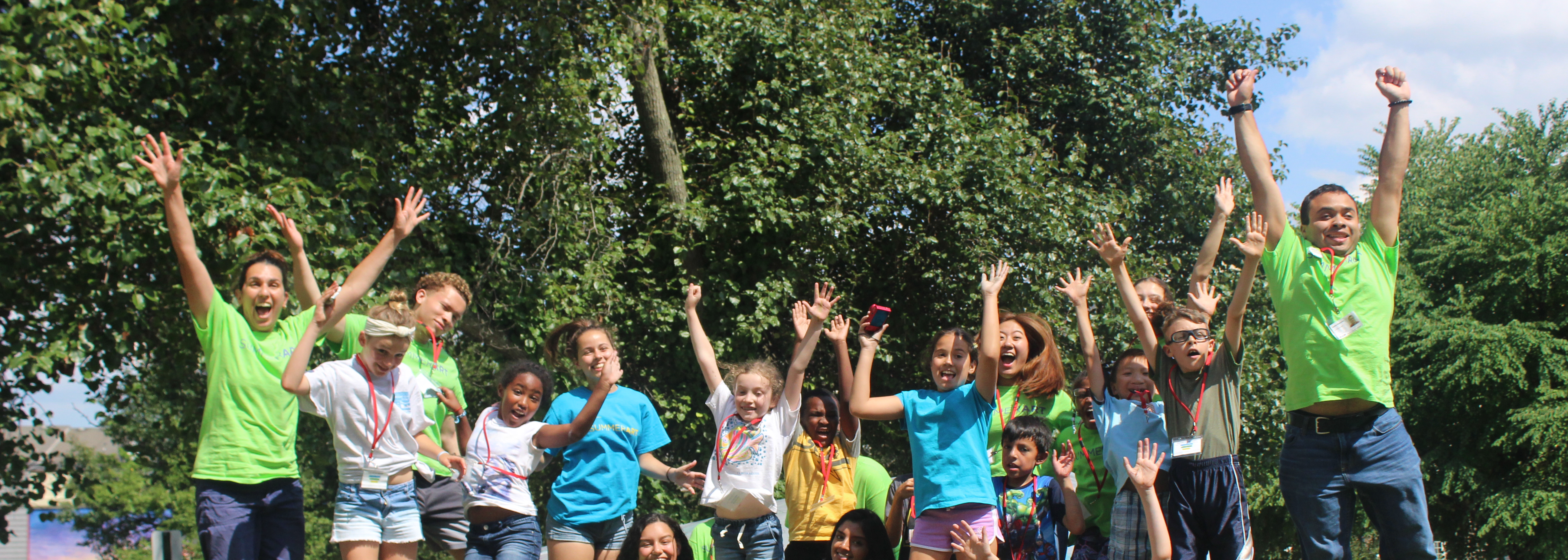 Summer art campers and counselors are all jumping and smiling at the camera with their hands in the air.