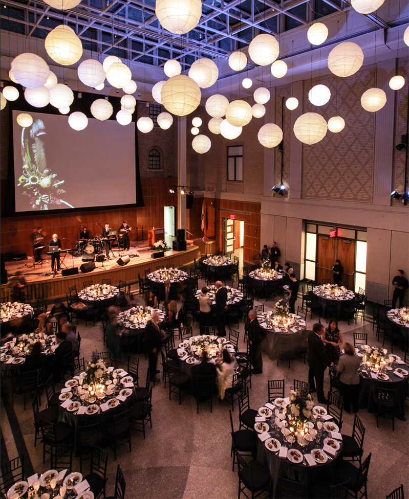 Bird eye view of Leir hall decorated with white hanging lanterns on the ceiling, formal dining tables, and a band on stage