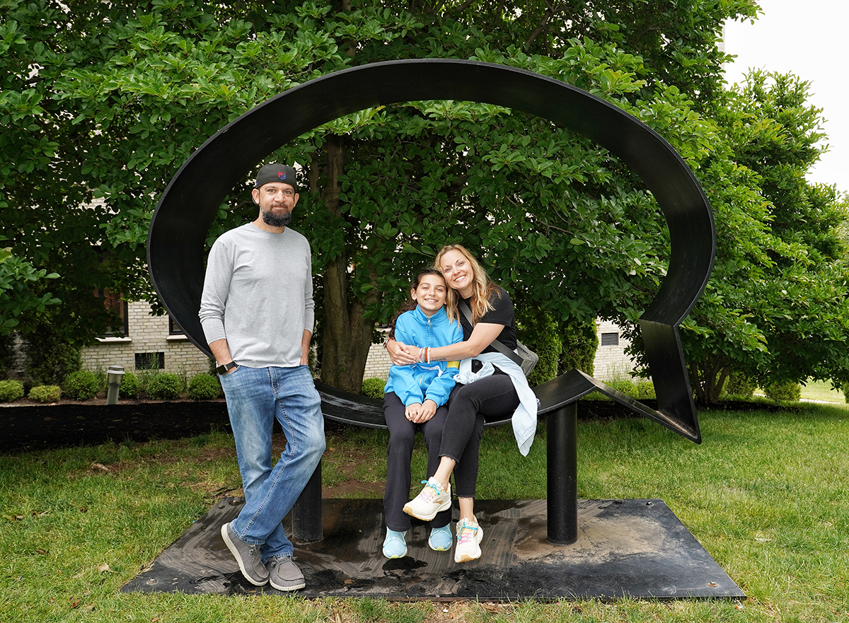 Family of three sitting in MAM's outdoor sculpture, "Speech Bubble", facing the camera and smiling