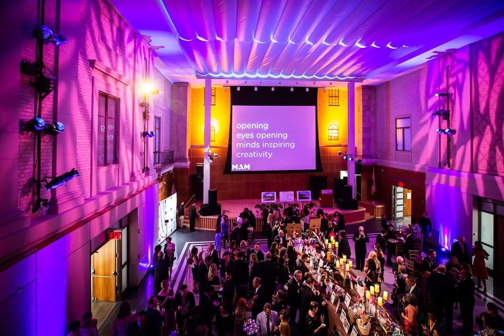 Gala event in a hall with colorful lighting, guests mingling, and a projector screen with text.