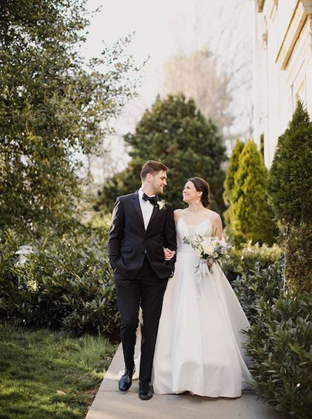A couple in wedding attire holding hands and walking down a garden path.