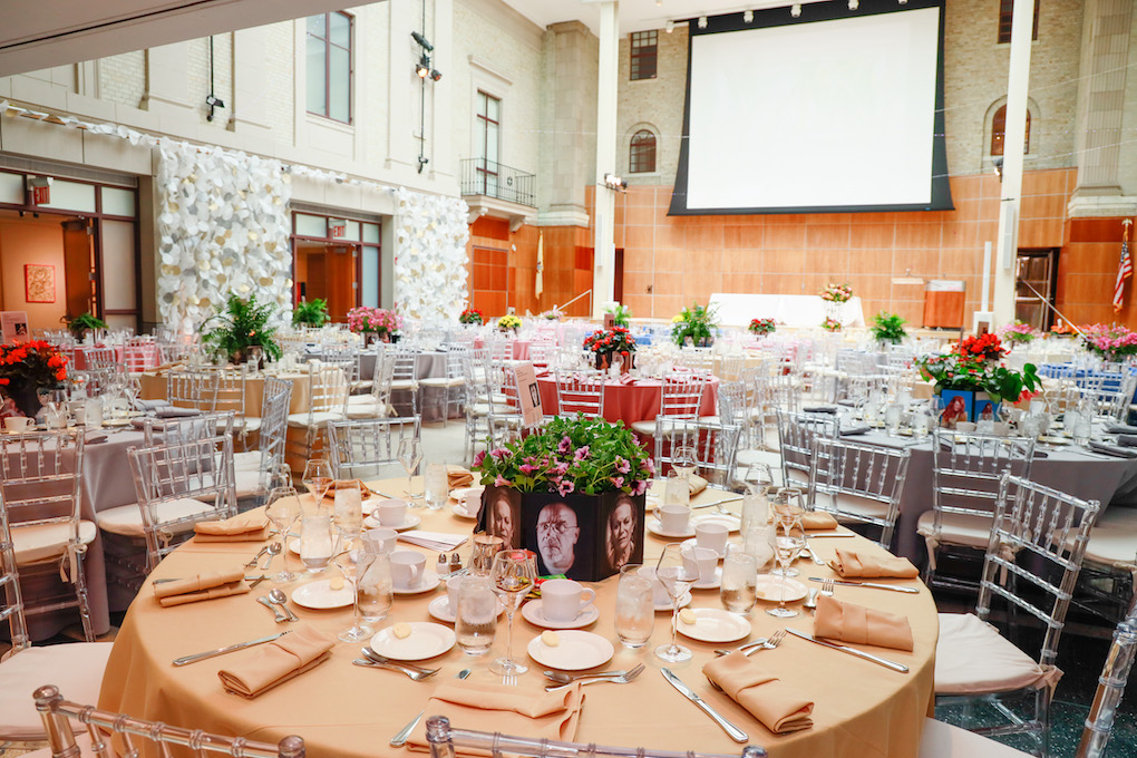 Elegantly set tables with clear chairs for an event in a spacious room with a large screen at the front.