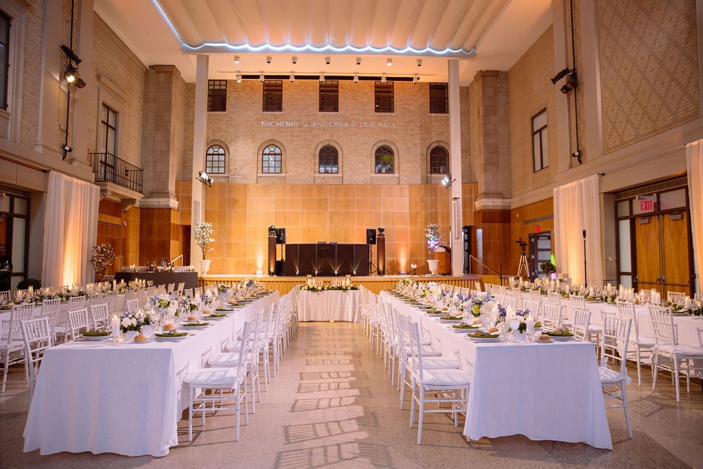 Elegant banquet setup in a spacious hall with white tables, chairs, floral centerpieces, and warm lighting.