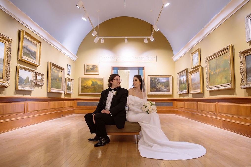 A couple in formal attire sits back-to-back on a bench in an art gallery, surrounded by various framed paintings on the wall.
