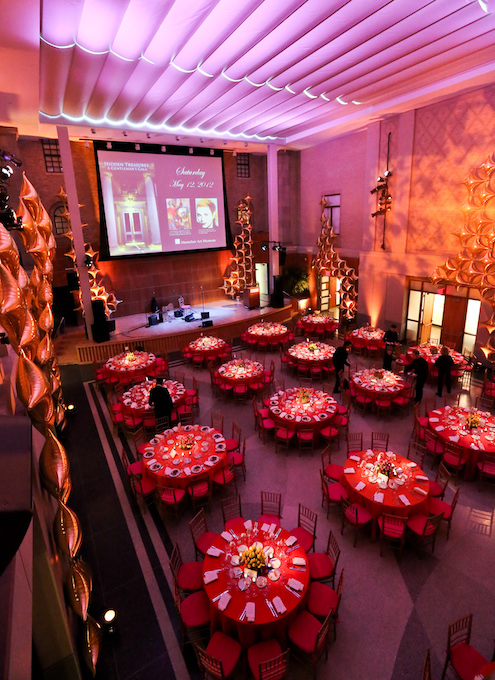Elegant banquet hall with red-themed tables, golden decor, and a stage displaying an event announcement under soft purple lighting.