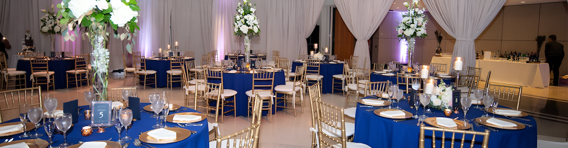 Elegant event space featuring gold chairs and blue tablecloths, adorned with floral centerpieces, candles, and a well-stocked bar.