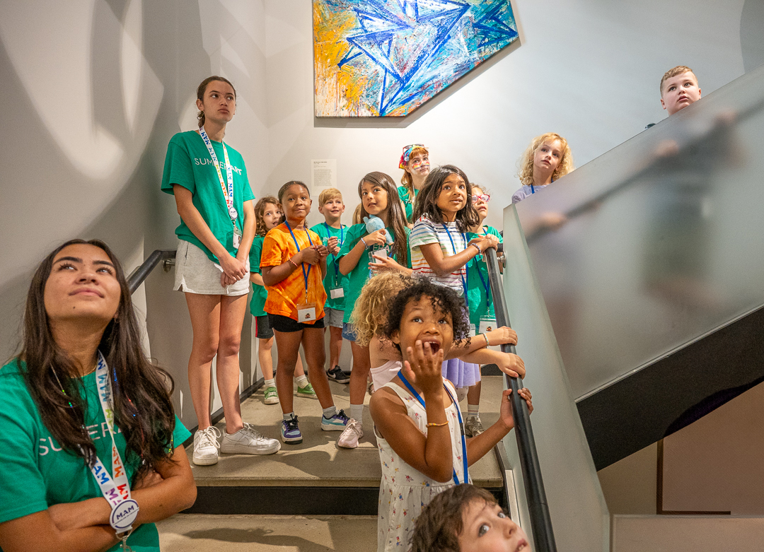 A group of children in colorful shirts and lanyards attentively observing art in a bright, modern gallery space.