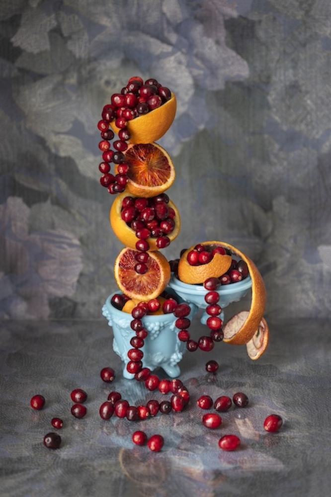 Colorful arrangement of sliced oranges and cranberries cascading from a blue bowl onto a textured background.