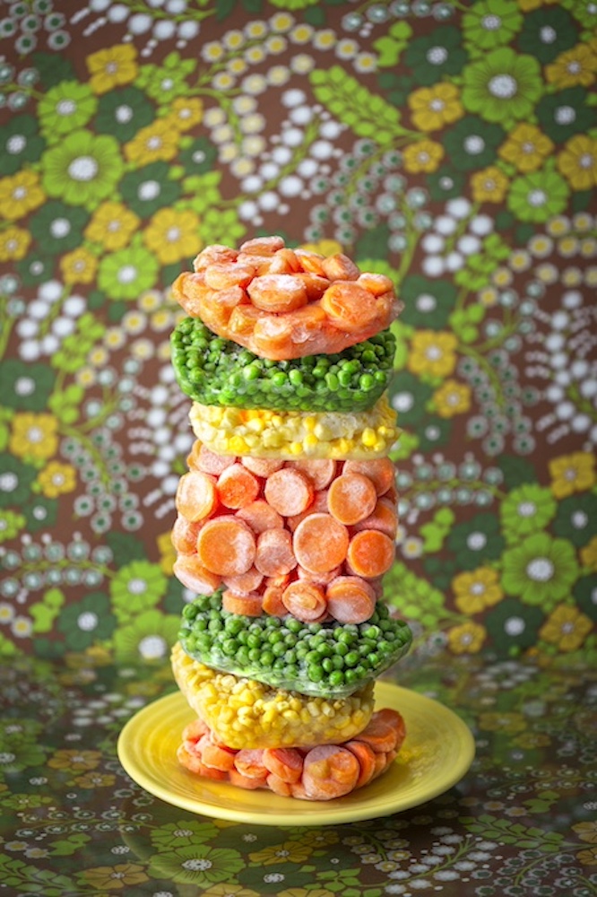 A vibrant stack of frozen vegetables, including carrots, peas, and corn, arranged on a yellow plate against a floral patterned background.