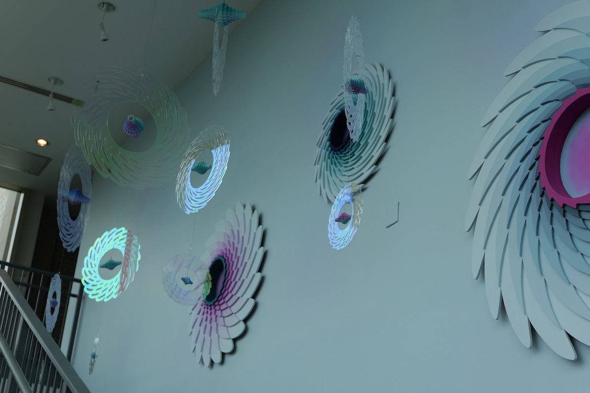 Colorful, layered paper sculptures resembling flowers or spirals hang gracefully against a pale wall, creating a dynamic, artistic installation.