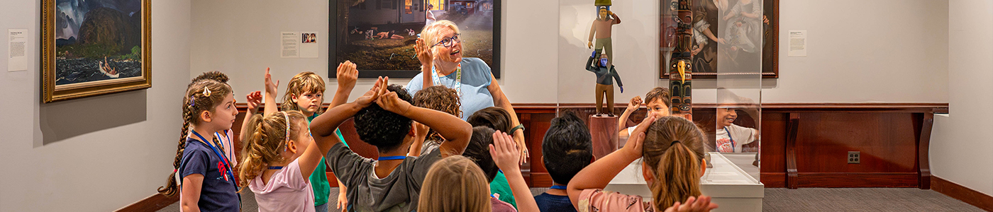 A group of excited children in a museum engaged in a learning activity, with artwork displayed in the background.