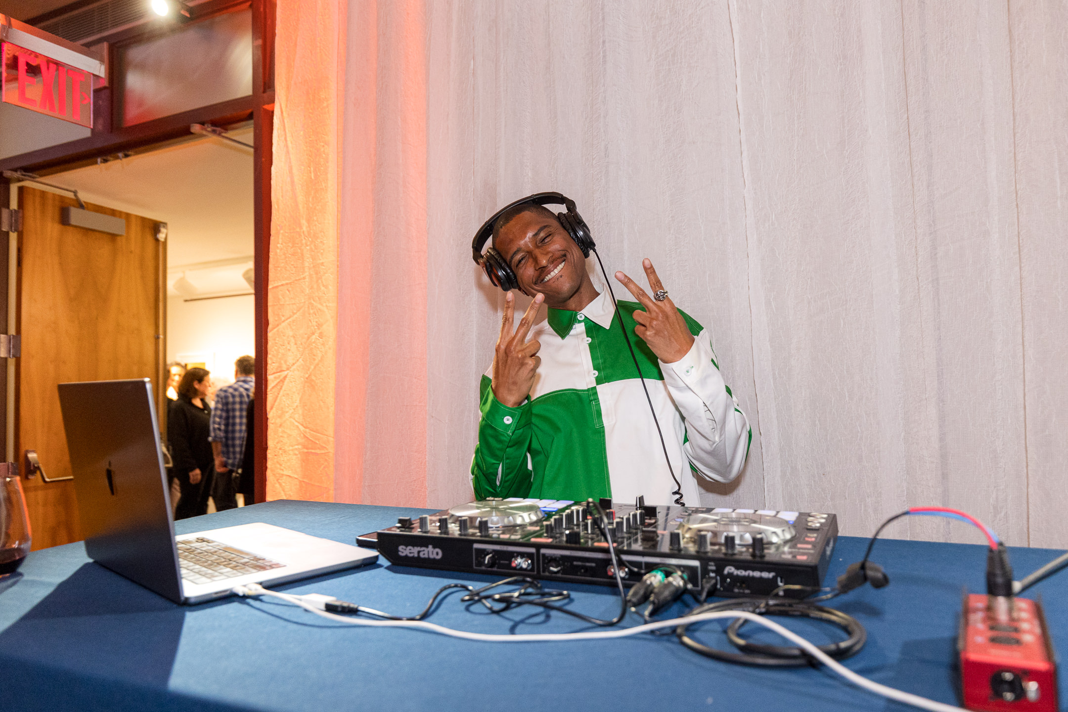 A DJ in a green and white shirt poses with peace signs, surrounded by a laptop and DJ equipment at a lively event.