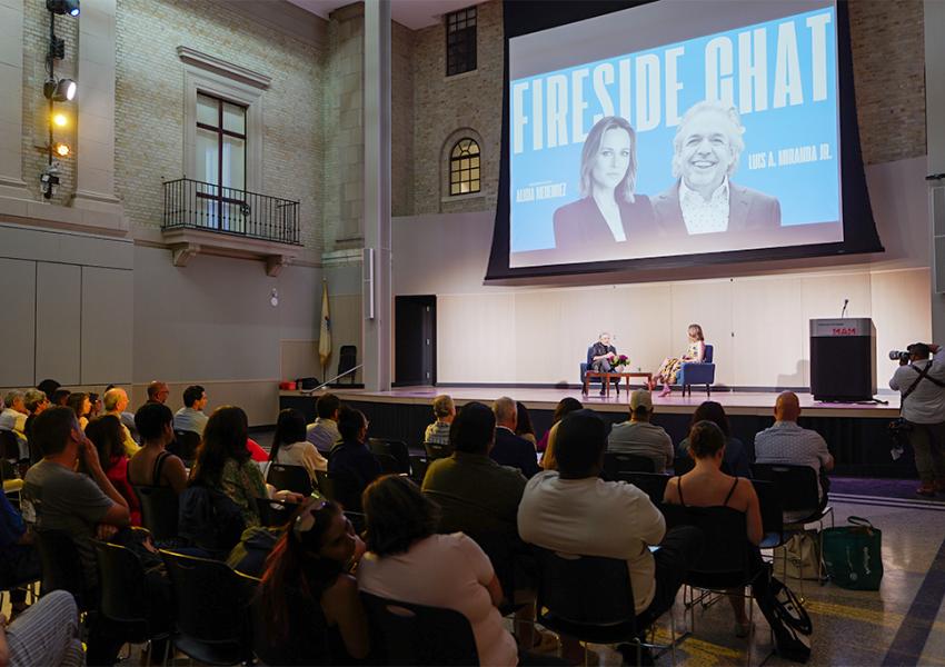 Audience watching a presentation with two speakers on stage and a "Fireside Chat" screen backdrop.