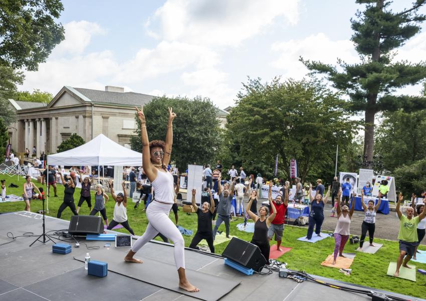 A yoga instructor leads a class outdoors, surrounded by participants on mats, with a historic building and tents in the background.