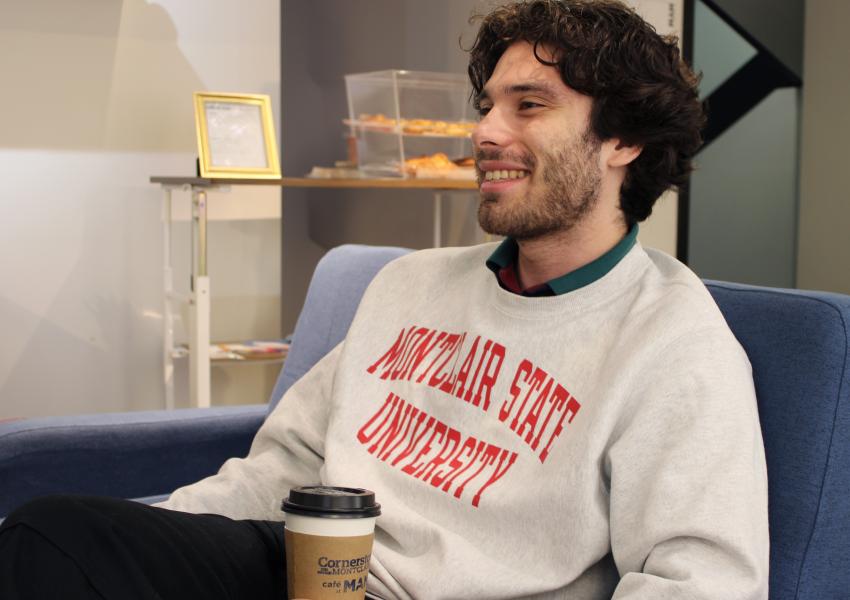 Student wear a "Montclair State University" sweatshirt, sitting with a to go coffee cup in their hand
