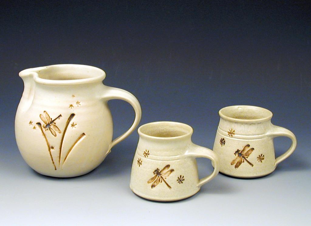 A creme colored pitcher and two mugs, created by hand on the pottery wheel. They've been set up on a backdrop for professional photography. 