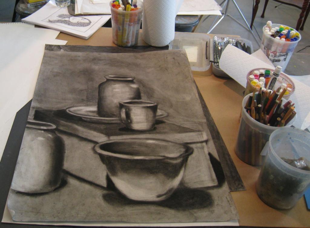 A student's workspace on a table in the studio from the view of the student. A Large sketchbook is in front of us, with a charcoal drawing of some bowls and bottles. To the right is a couple of cups full of pencils and other drawing utensils.