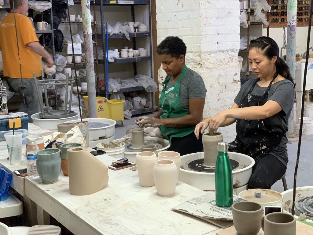 two students are working on the pottery wheels in the ceramic studio. Both are focused on their clay cylinders and not looking at the camera. Instructor Steve is in the background, looking a finished pieces on the shelves. 