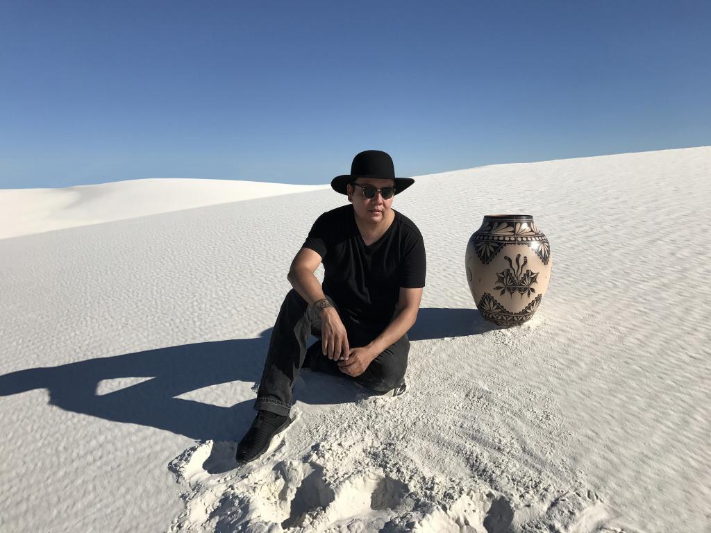 Virgil ortiz is sitting on a sand dune next to one of his elaborately decorated pots that is 1.5-2 feet tall.