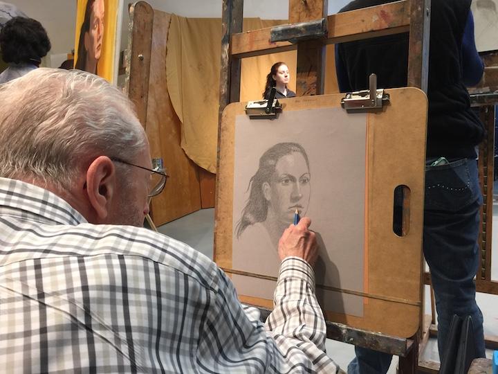A student is sitting at an easel, drawing a portrait of a woman with a pencil.