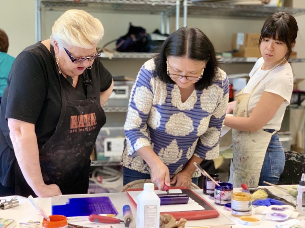 A woman is rolling ink onto a breyer with dorothy cochran standing next to her and looking on. Another student is behind them watching.