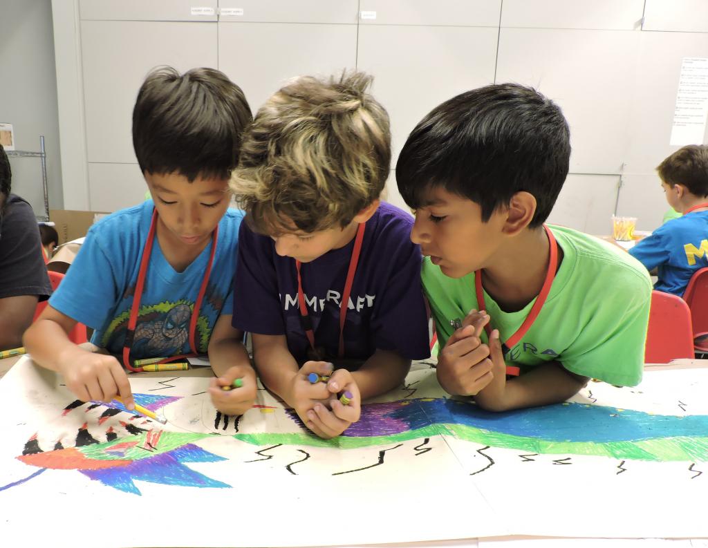 Three campers are working together on a drawing of a dragon on a table.