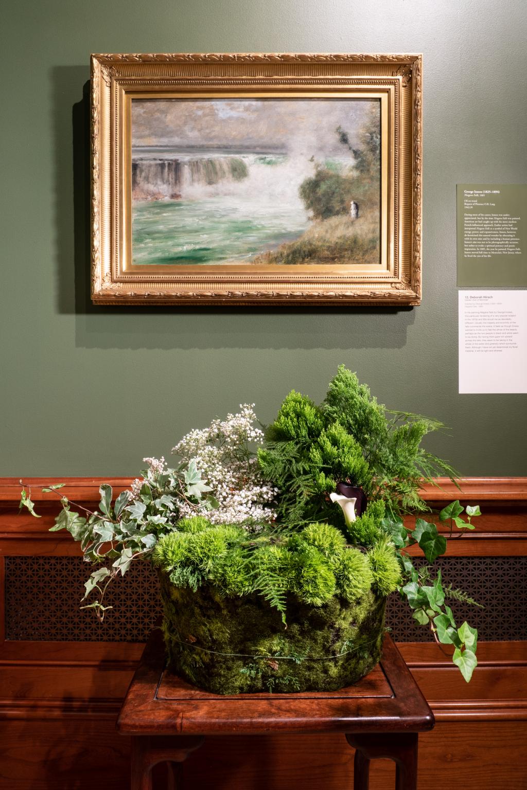 a wide, green floral arrangement is sitting on a wooden end table underneath an inness painting of a waterfall.