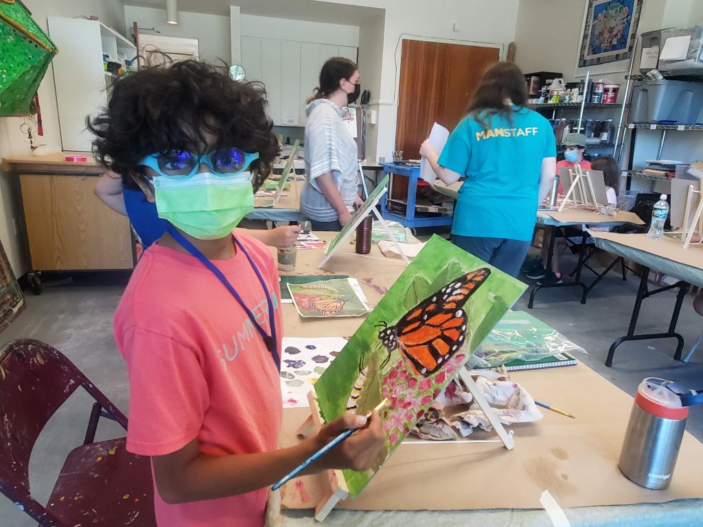 A young student is wearing a mask and looking at the camera while painting a work on canvas.