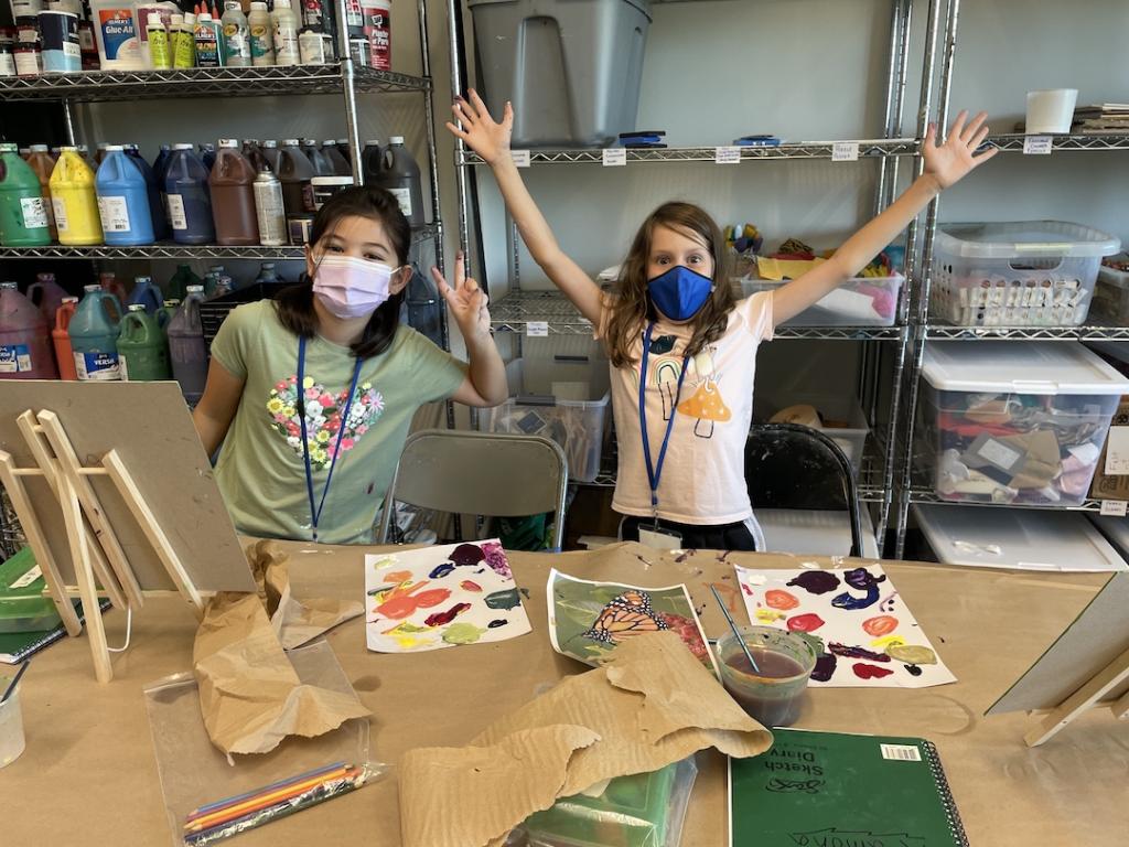 Two young students posing for the camera during painting camp with materials strewn in front of them.