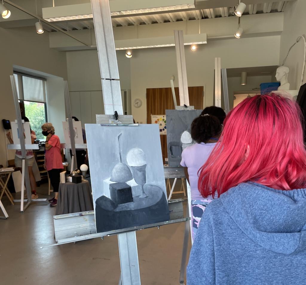 Looking over the should of a student with bright red hair at their greyscale still life painting in progress on an easel.