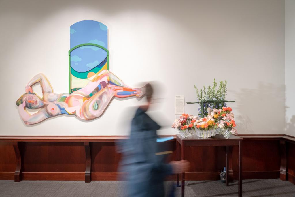 A visitor appears blurry as they walk in front of an artwork and a floral arrangement.