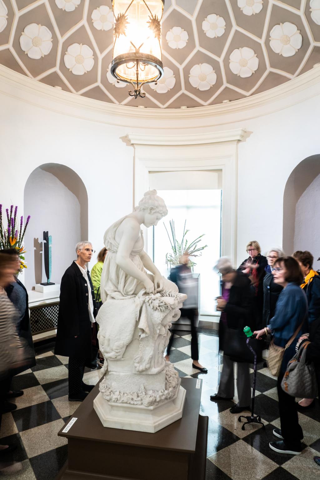 A tour group in the Rotunda gallery looking at the Crown for the Victor sculpture with a floral arrangement behind it.