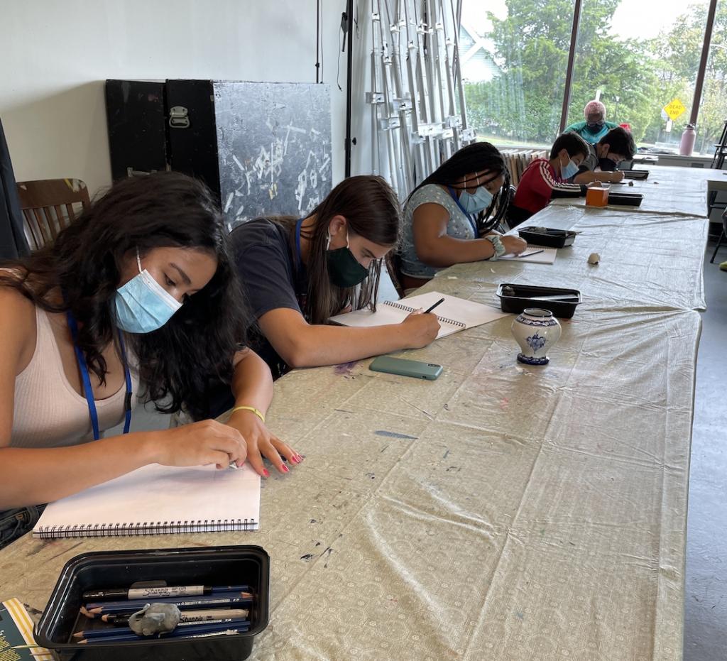 A row of teen students drawing sketchbooks at a long table.