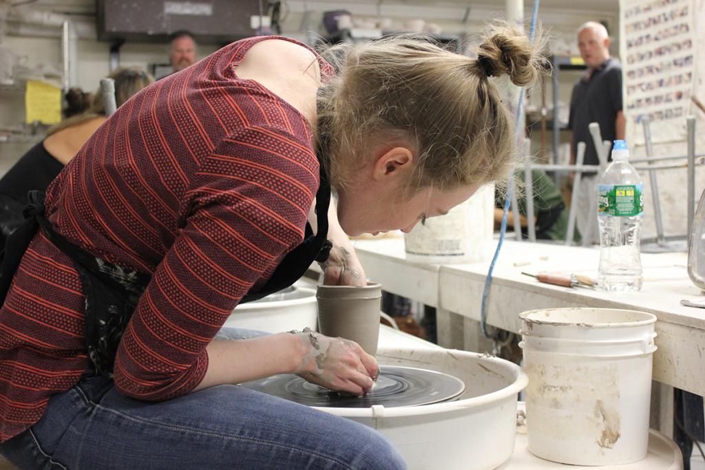 Beginner Wheel Throwing: View from the side as a student sits at their potter's wheel. They are in the middle of throwing a tall vase, one hand is inside the vase, while the other holds the vessel steady on the outside.
