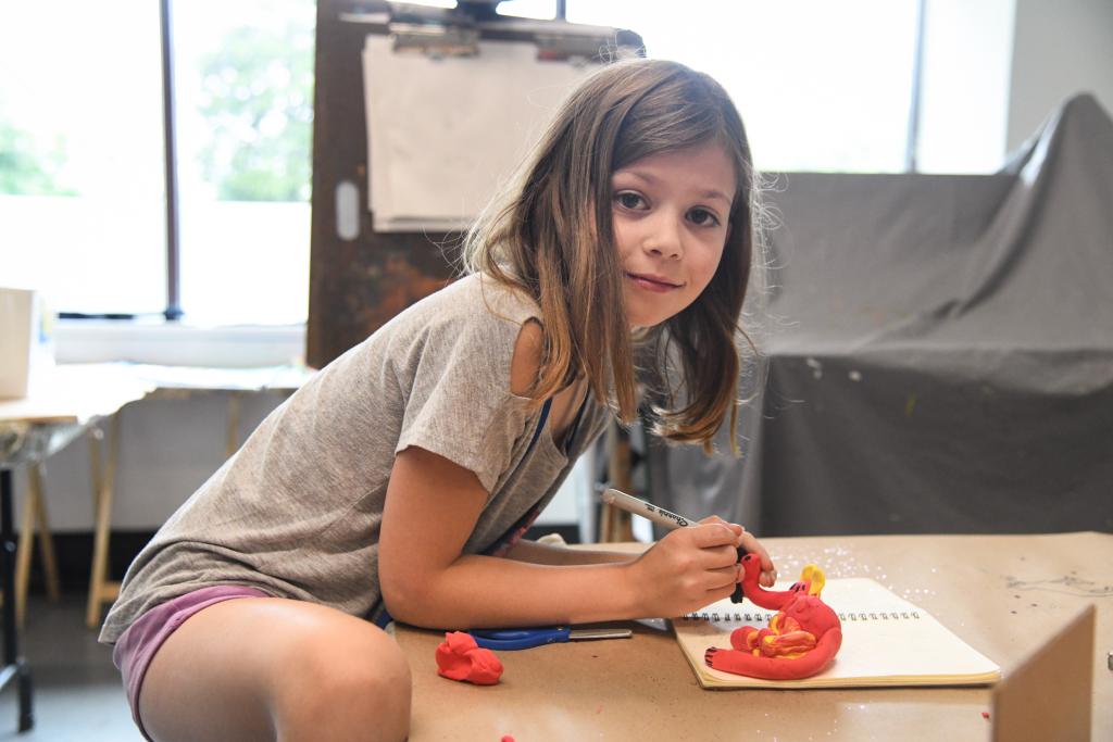 Side facing view of young student hunched over a table as they craft a figurine out of red and orange quick dry clay