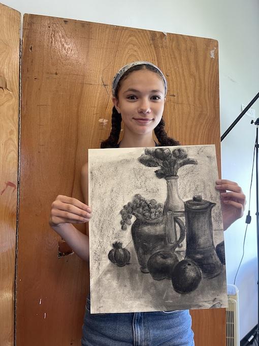 Teen student holding up a charcoal still life drawing containing a vase of flowers, a pitcher, grapes, an apple, and an orange