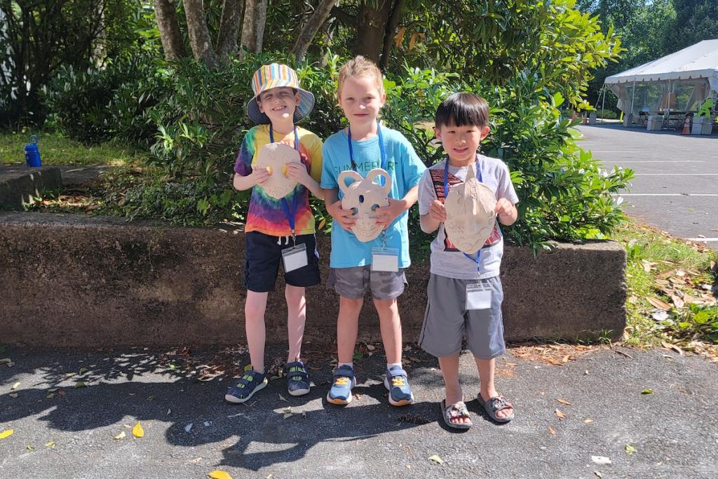 Three children holding paper masks, standing on a sidewalk with greenery in the background.