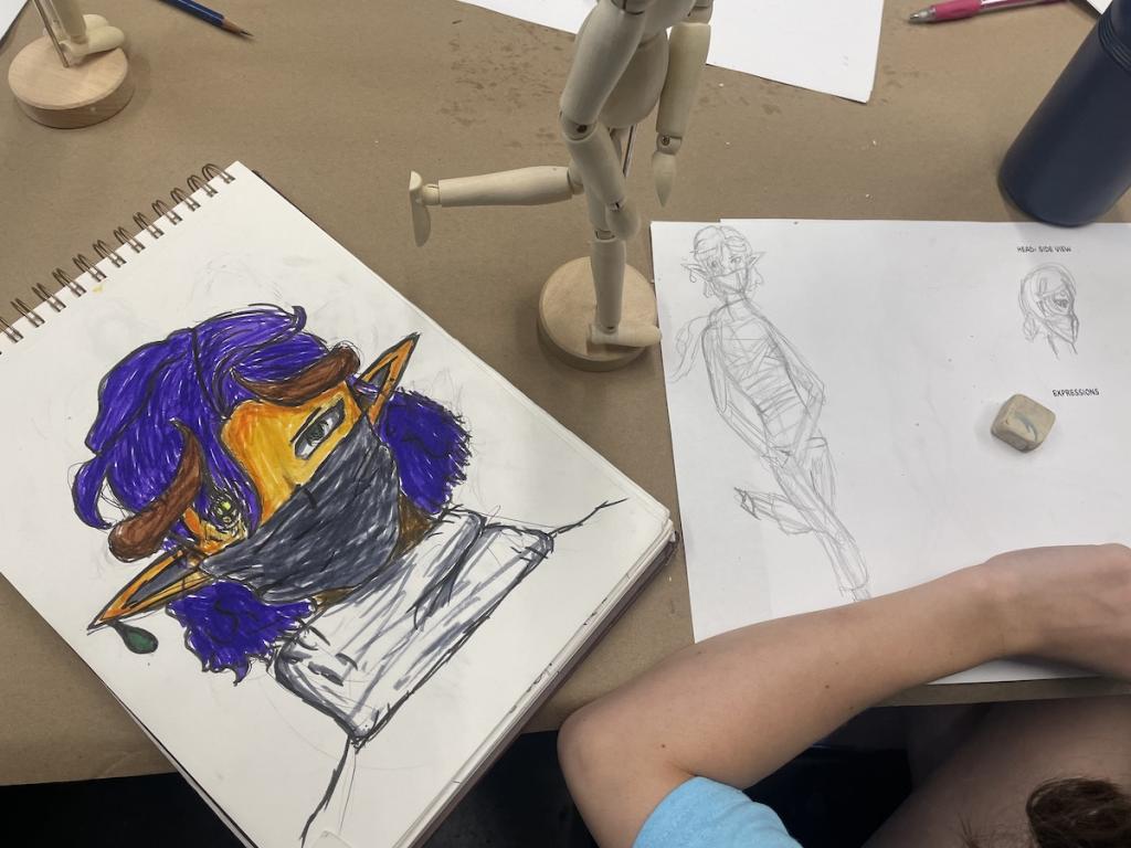 Sketches on paper, one with a colorful stylized character and another with a human figure, beside a wooden mannequin.