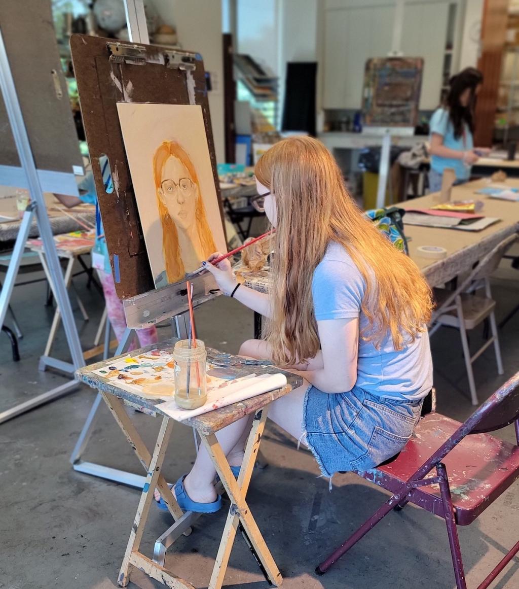 Girl painting on canvas in an art studio, surrounded by art supplies.