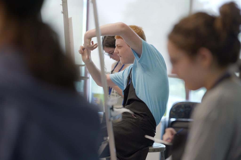 A person in a blue shirt painting at an easel in a bright classroom.