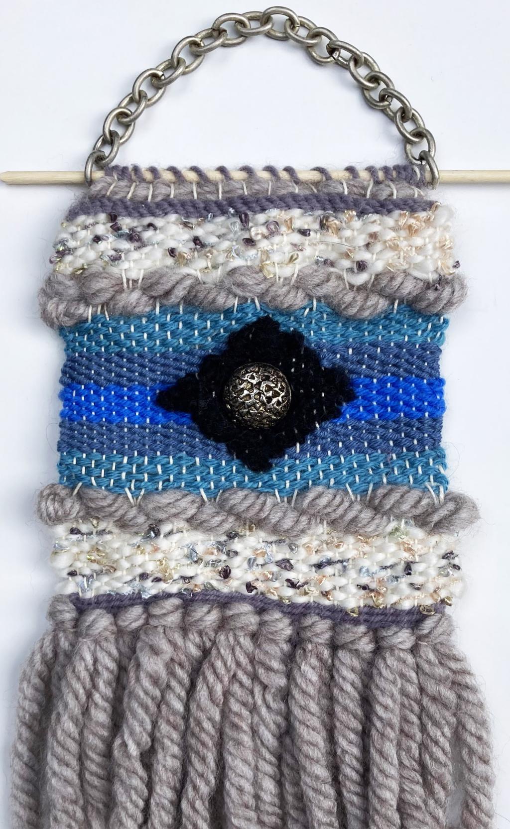 A handmade textile wall hanging with varying shades of blue, cream, and gray, featuring a central black pattern and decorative fringe.