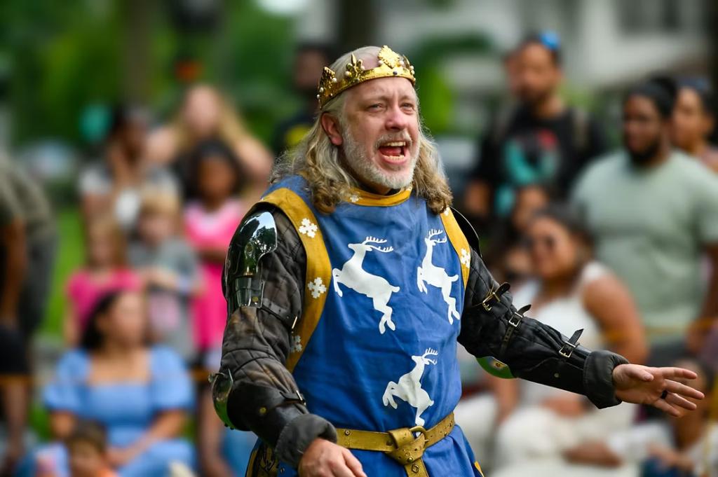 A man in medieval armor and a blue tunic with white deer motifs performs in front of an engaged audience at an outdoor event.